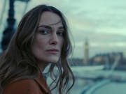 A white woman with long brown hair wearing a brown jacket looking serious and windswept standing beside the River Thames with Big Ben in the background