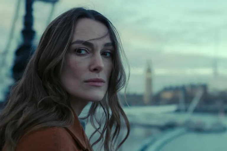 A white woman with long brown hair wearing a brown jacket looking serious and windswept standing beside the River Thames with Big Ben in the background