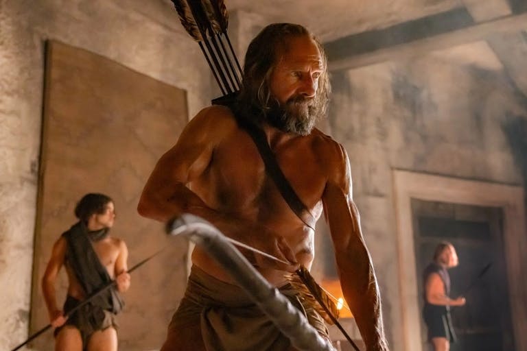 A topless strong middle aged man holding a bow with arrows strapped to his back with two younger men in the background also with bows