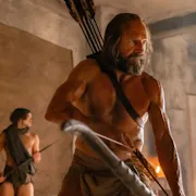 A topless strong middle aged man holding a bow with arrows strapped to his back with two younger men in the background also with bows