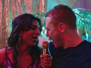 A young Asian drag queen in a sparkly strappy dress singing into a gold mic with a young white man in a black top