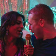 A young Asian drag queen in a sparkly strappy dress singing into a gold mic with a young white man in a black top