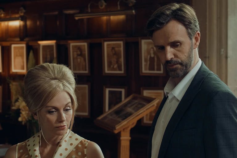 A white woman with bouffant blonde hair standing next to a white man in a suit jacket and white shirt, looking serious