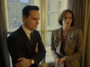 A white man in a dark suit and a white woman in a light suit stand at a window looking thoughtful