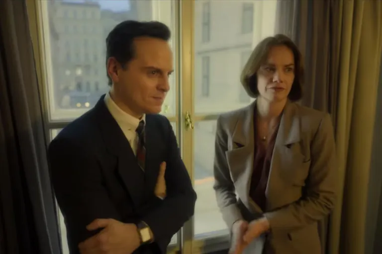 A white man in a dark suit and a white woman in a light suit stand at a window looking thoughtful