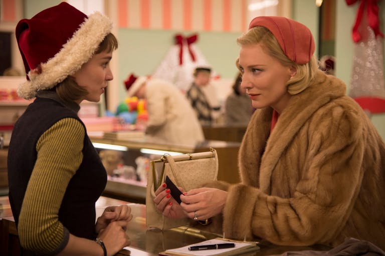 A young white woman with short brown hair wearing a velvet Santa hat stands across the counter from an older white woman with short blonde hair wearing a light fur coat