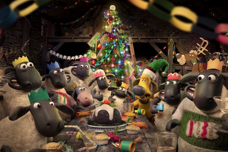 A group of stop motion plasticine sheep wearing Christmas cracker hats gathered around a table of food with a smaller sheep at the head of the table alongside a light brown dog, a twinkling Christmas tree in the background