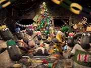 A group of stop motion plasticine sheep wearing Christmas cracker hats gathered around a table of food with a smaller sheep at the head of the table alongside a light brown dog, a twinkling Christmas tree in the background