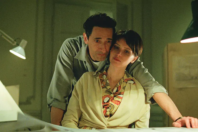 A white man leaning over a white woman his arm around her, looking at something on a desk
