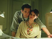 A white man leaning over a white woman his arm around her, looking at something on a desk
