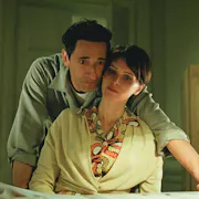 A white man leaning over a white woman his arm around her, looking at something on a desk