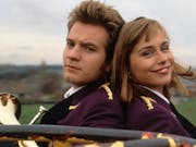 White man and white woman sitting back to back facing the camera. The man is holding a brass instrument