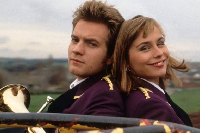 White man and white woman sitting back to back facing the camera. The man is holding a brass instrument