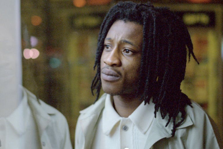 Black man with dreadlocks and  white t-shirt and cream coat, is looking out of window