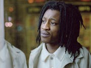 Black man with dreadlocks and white t-shirt and cream coat, is looking out of window