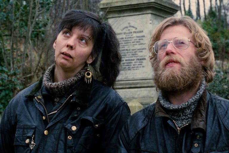 A blonde man with a beard wearing glasses and woman with black hair in a highponytail are looking curiously in the distance