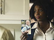 Black woman with afro hair, wearing a white shirt and black cardigan is showing the picture of a young black girl to someone.