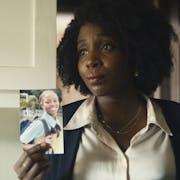 Black woman with afro hair, wearing a white shirt and black cardigan is showing the picture of a young black girl to someone.