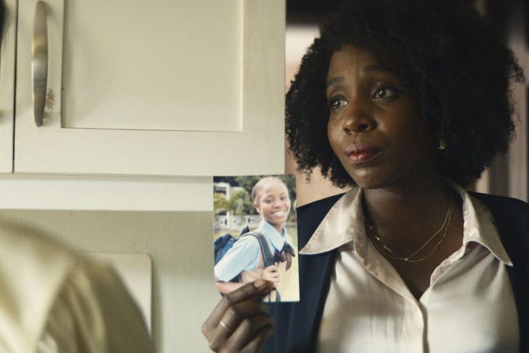Black woman with afro hair, wearing a white shirt and black cardigan is showing the picture of a young black girl to someone.