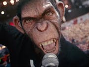 A monkey, looking at the camera is holding a microphone and singing. In the background a large concert crowd