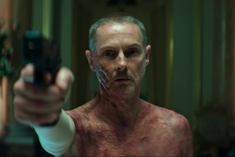 A white man with short grey hair and sever burns on his chest and face is pointing a gun towards the left of the camera