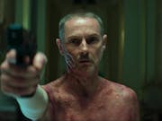 A white man with short grey hair and sever burns on his chest and face is pointing a gun towards the left of the camera