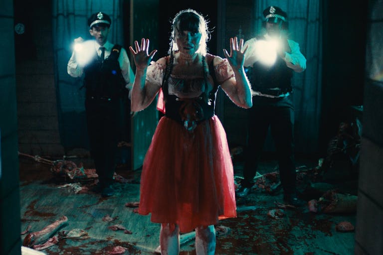 A woman in red dress facing the camera is covered in blood and shows us her hands, behind her wo police officers with flashlights point them at our direction.