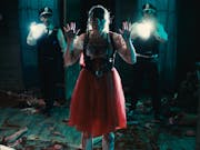 A woman in red dress facing the camera is covered in blood and shows us her hands, behind her wo police officers with flashlights point them at our direction.