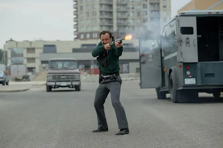 A white man is holding a shotgun and shoots towards the right of the camera. He is near a van and in the background, a highrise building