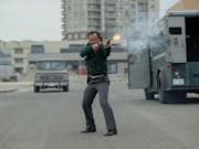 A white man is holding a shotgun and shoots towards the right of the camera. He is near a van and in the background, a highrise building