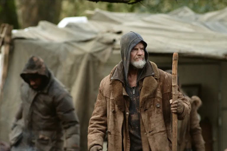 A white man holding a wooden staff, is wearing a thing jacket and a hoodie is standing in front of a makeshift tent. Thwo other people in hooded jackets are in the background