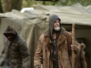 A white man holding a wooden staff, is wearing a thing jacket and a hoodie is standing in front of a makeshift tent. Thwo other people in hooded jackets are in the background