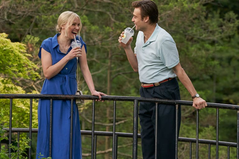 A white man and a white woman in a blue dress standing on a bridge are drinking for plastics cups and laughing looking at each other. Dense foliage in the background