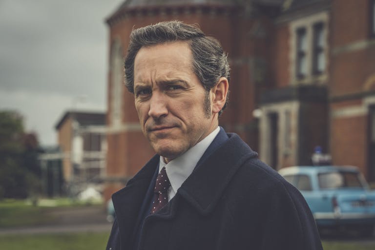 A white man wearing a suit and red tie is standing in front of red brick building looking at the camera at a 3/4 angle