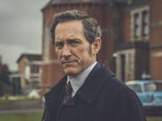 A white man wearing a suit and red tie is standing in front of red brick building looking at the camera at a 3/4 angle