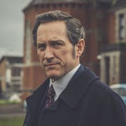 A white man wearing a suit and red tie is standing in front of red brick building looking at the camera at a 3/4 angle