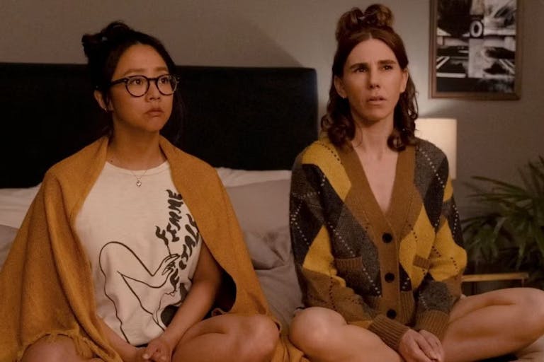 An Asian woman and a white woman sitting on a bed with their legs crossed looking at something off screen