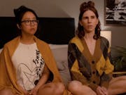 An Asian woman and a white woman sitting on a bed with their legs crossed looking at something off screen