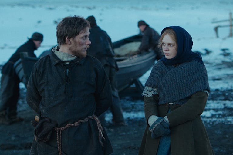 A young white man and woman in plain 19th Century clothing walking along a snowy shore, with two men in the background dragging a small boat