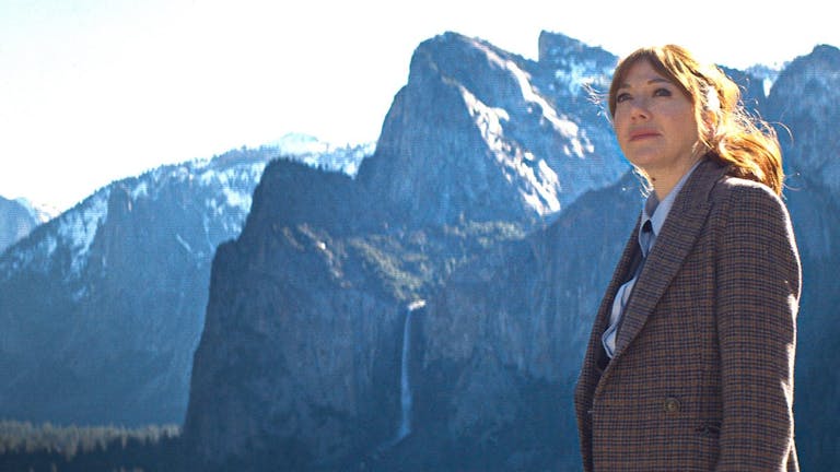 A white woman with red hair in a suit looking contemplative with dramatic snowy topped mountains in the background