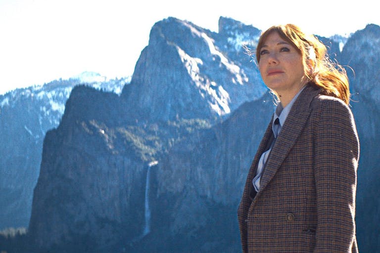 A white woman with red hair in a suit looking contemplative with dramatic snowy topped mountains in the background