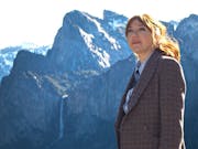 A white woman with red hair in a suit looking contemplative with dramatic snowy topped mountains in the background