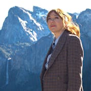 A white woman with red hair in a suit looking contemplative with dramatic snowy topped mountains in the background