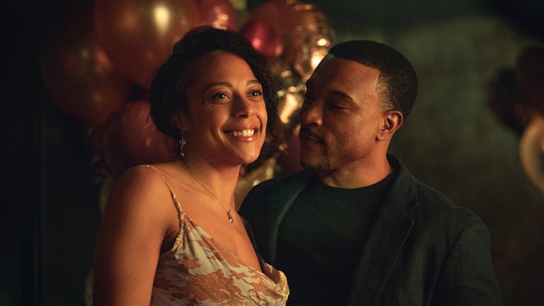 A Black woman in a cocktail dress and a Black man in a black suit at a party looking happy