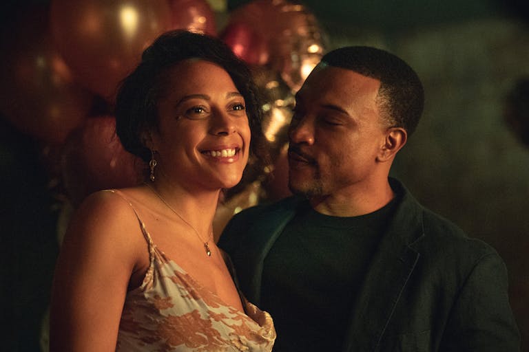 A Black woman in a cocktail dress and a Black man in a black suit at a party looking happy