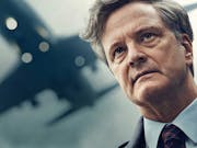A middle aged white man with grey hair wearing a suit looking haunted as a plane flies overhead