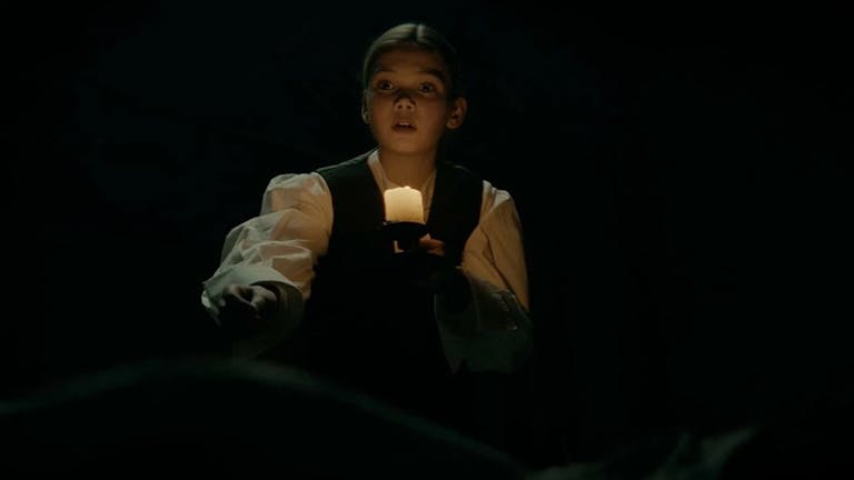 A young white girl in period clothes, in the dark holding a lamp illuminating her face