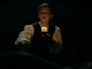 A young white girl in period clothes, in the dark holding a lamp illuminating her face