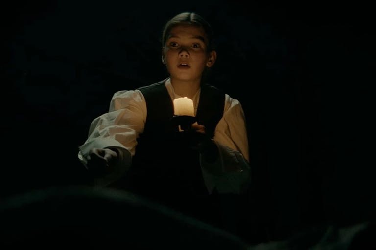 A young white girl in period clothes, in the dark holding a lamp illuminating her face