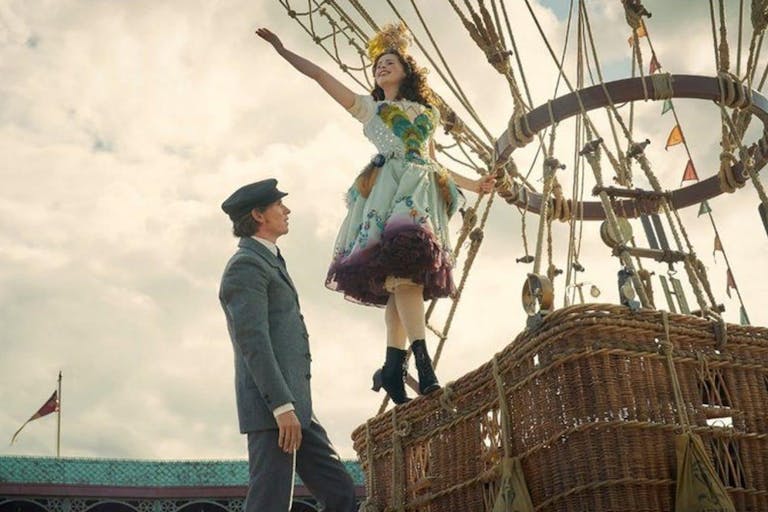 A young white man in a suit stands at the foot of a hot air balloon as a young white woman in a colourful dress stands on the basket with her arm in the air